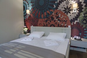 A hotel room with a bed, two bedside tables, a wall with a colorful geometric pattern, and a vase with flowers on one of the tables. - Zepter Hotel Drina (Bajina Bašta)