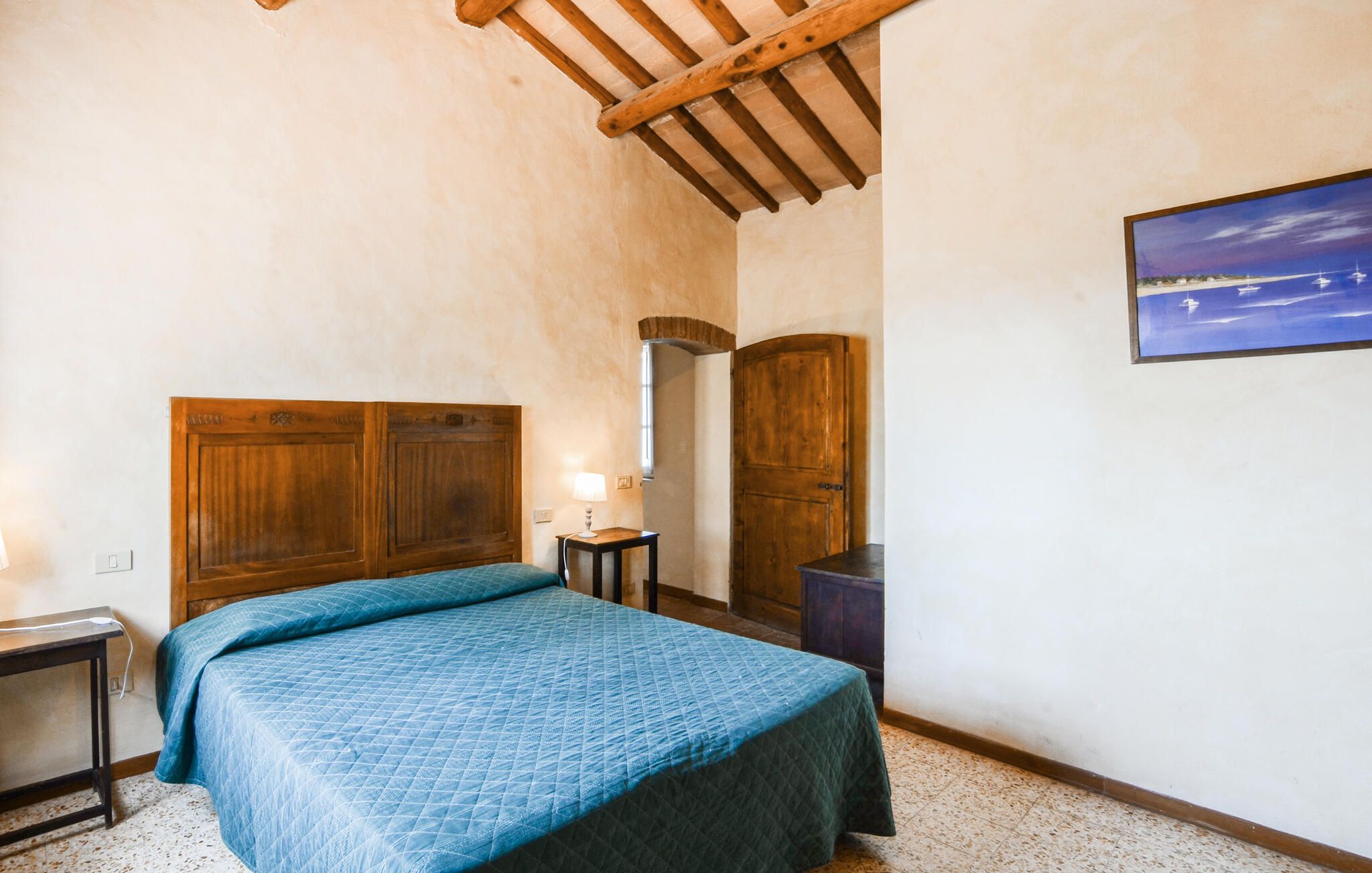 A bedroom with a wooden headboard, a bed with a blue quilt, a nightstand, a small table with a lamp, a wooden door, and a framed picture on the wall.