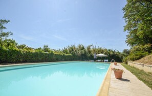 Ein Schwimmbecken mit Holzterrasse, umgeben von viel Grün und blauem Himmel.