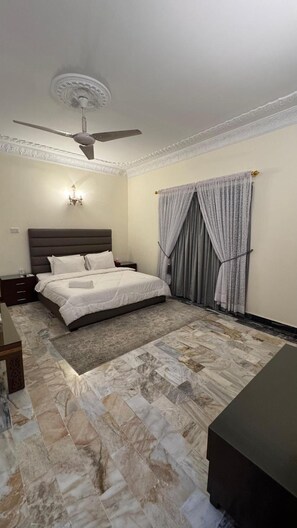 Une chambre à coucher avec un lit, un ventilateur de plafond, un lustre, des rideaux et un sol à motifs de marbre.