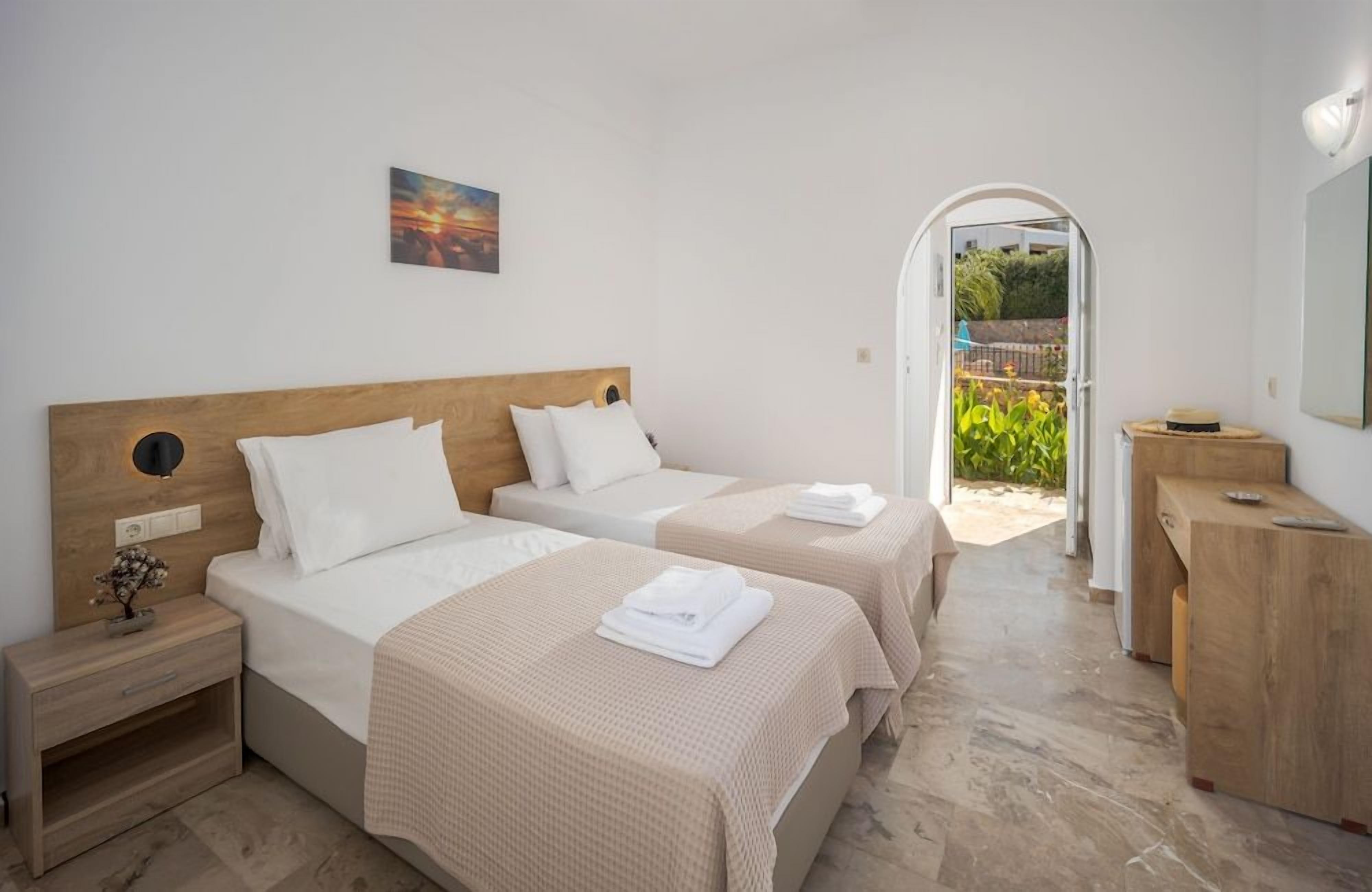A hotel room with two beds, a wooden headboard, a bedside table, a nightstand, a mirror, and a view of the garden through an open door.