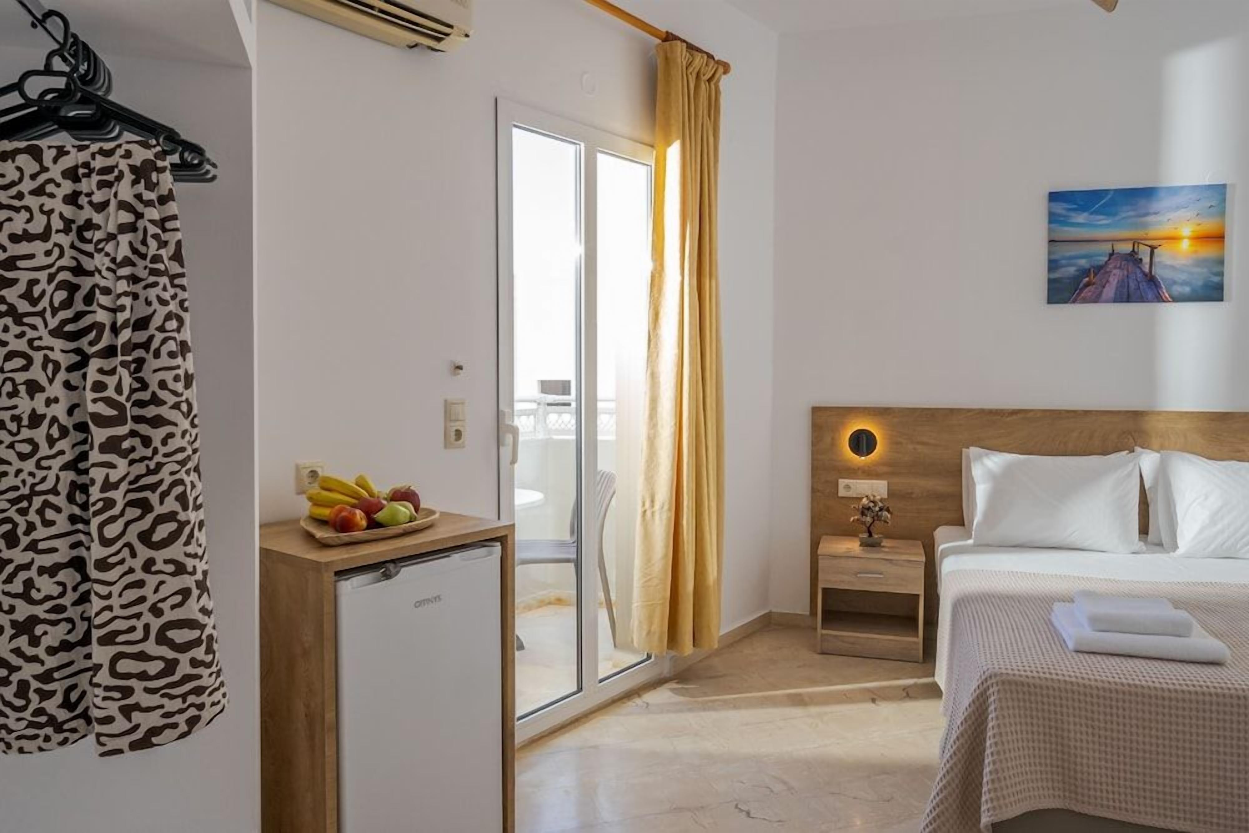 A hotel room with a bed, a bedside table, a small desk with a refrigerator, a window with curtains, and a painting on the wall.