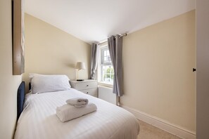 Una cama bien hecha con ropa blanca y una toalla doblada, una lámpara de noche, una ventana con cortinas y un radiador.