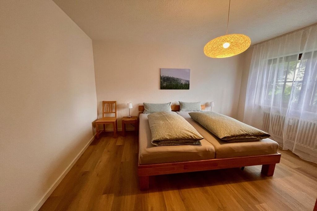 A bedroom with a wooden bed, a bedside table, a chair, and a hanging light fixture.