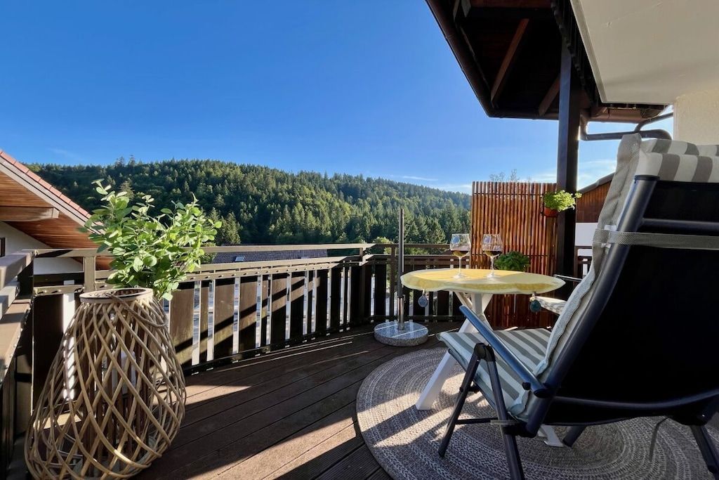 A wooden deck with a table and chairs, a hanging plant, and a view of a forested hill.