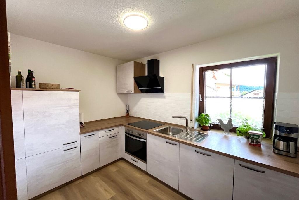 Une cuisine moderne avec des meubles blancs, un four encastré, un évier et une fenêtre donnant sur un beau paysage.