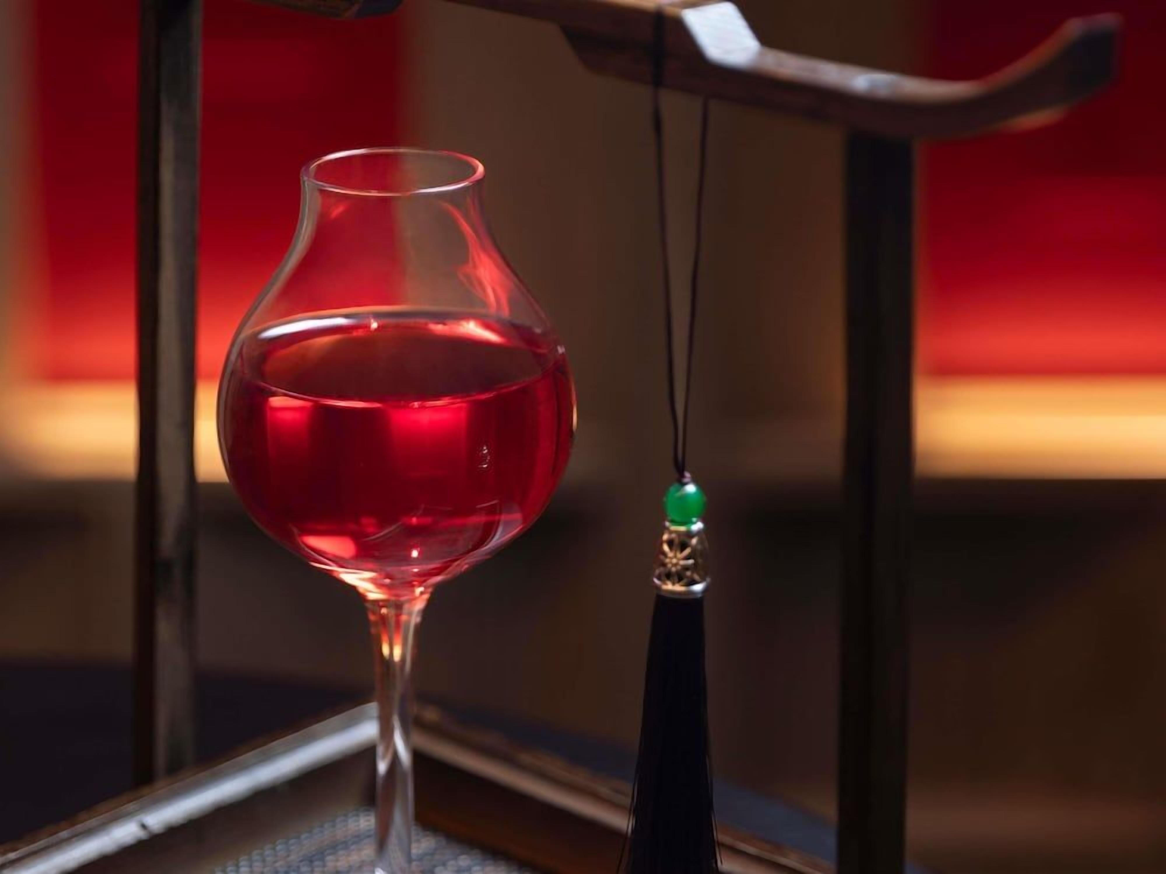 Una copa de vino tinto llena, sostenida por un hilo con una cuenta verde, sobre un fondo desenfocado.
