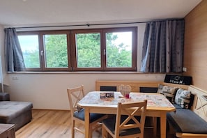 Ein gemütliches Zimmer mit Holztisch, Stühlen und einer Bank. Es gibt ein Fenster mit Vorhängen, einen kleinen Tisch mit einer Dekoration und ein an der Wand befestigtes Radio.
