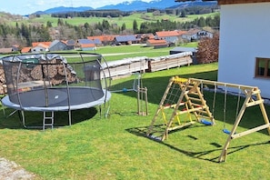 Ein wohnbereichsähnlicher Außenbereich mit Trampolin, Holzschaukel und Blick auf ein Dorf mit Häusern und Solaranlagen.