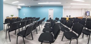 Una sala conferenze con file di sedie nere, un tavolo bianco con una pianta e uno schermo per presentazioni.