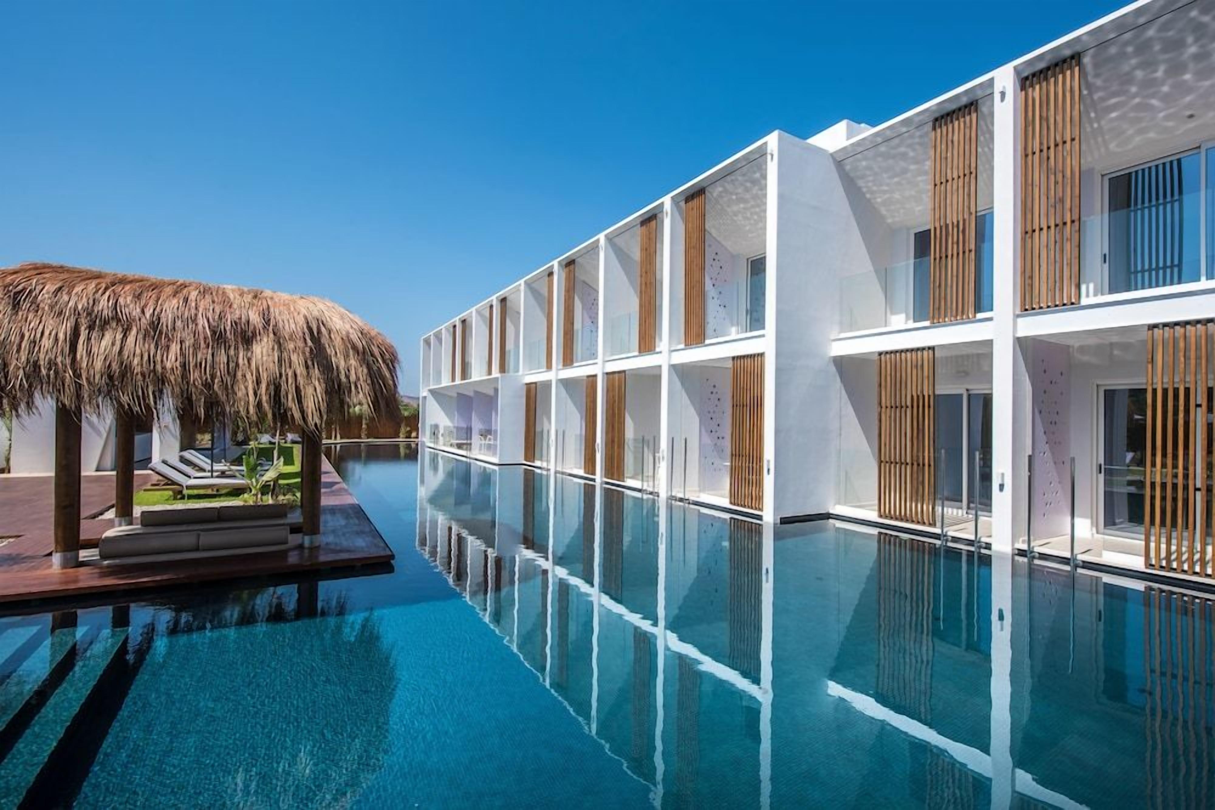 Un hôtel moderne doté d’une piscine à débordement en verre, d’éléments décoratifs en bois et d’une structure surmontée d’un toit de chaume.