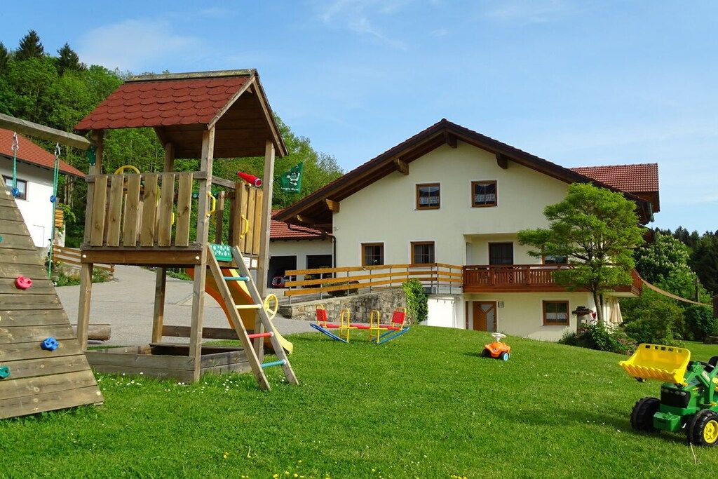 Un parque infantil con una estructura de madera para escalar y columpios, un tractor verde, y una casa con techo rojo y balcón.
