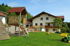 Ein Spielplatz mit einer hölzernen Kletterkonstruktion und Schaukeln, einem grünen Traktor und einem Haus mit rotem Dach und Balkon.