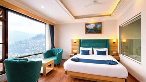 Ein Hotelzimmer mit einem großen Bett, einer Sitzecke inklusive Sessel und einem kleinen Tisch, sowie Blick auf eine Stadtkulisse durch große Fenster.