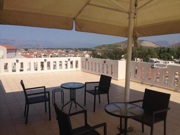 Uma área de estar ao ar livre com mesas e cadeiras, um grande guarda-sol e vista para uma cidade e colinas ao longe.