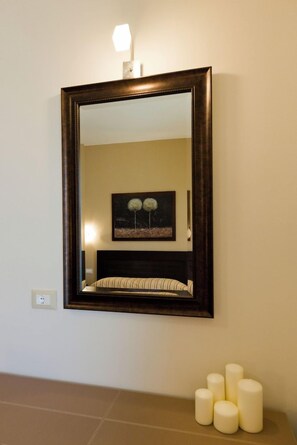 A mirror with a dark frame, a wall-mounted light, and a painting of trees. - Aenaon Apartments (Nafplio)