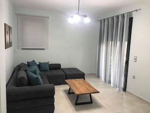 A living room with a sectional sofa, a wooden coffee table, a ceiling light fixture, and a window with curtains. - Aenaon Apartments (Nafplio)
