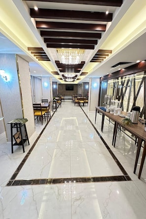 Corredor de hotel com candelabro, mesas com cadeiras e piso de mármore polido.