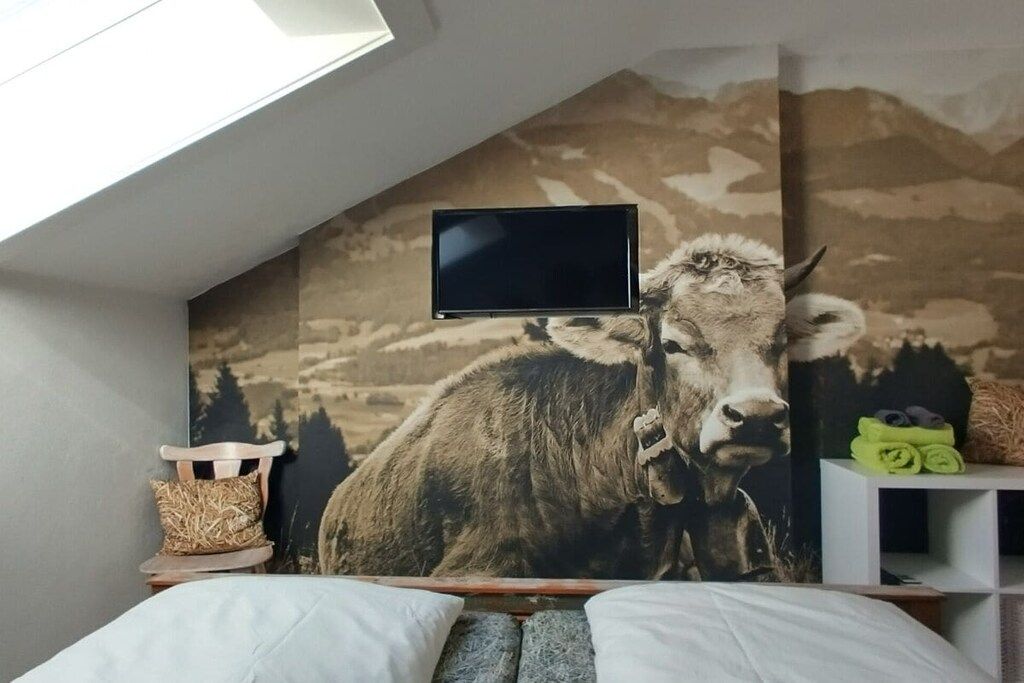 A bedroom with a mountain-themed wall, a mounted TV, a bed, a small wooden chair, and a shelf with towels.