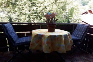 En terrasse med et rundt bord dækket af et blomstret dug, to blå polstrede stole og en potteplante med blomster.