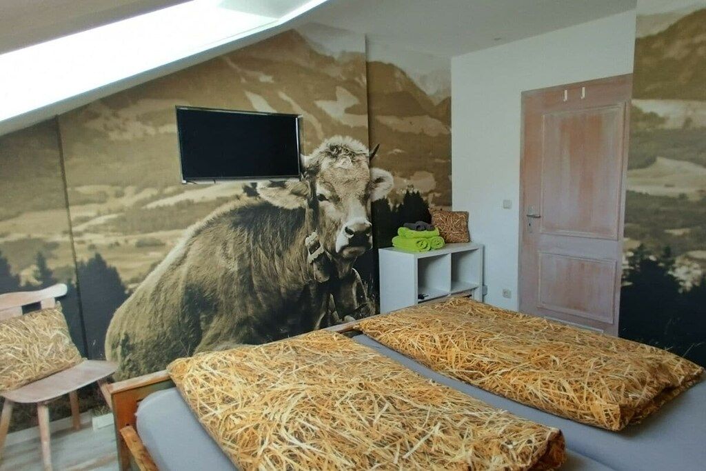 A room with a mural of a cow, a television mounted on the wall, and a bed with straw bedding.