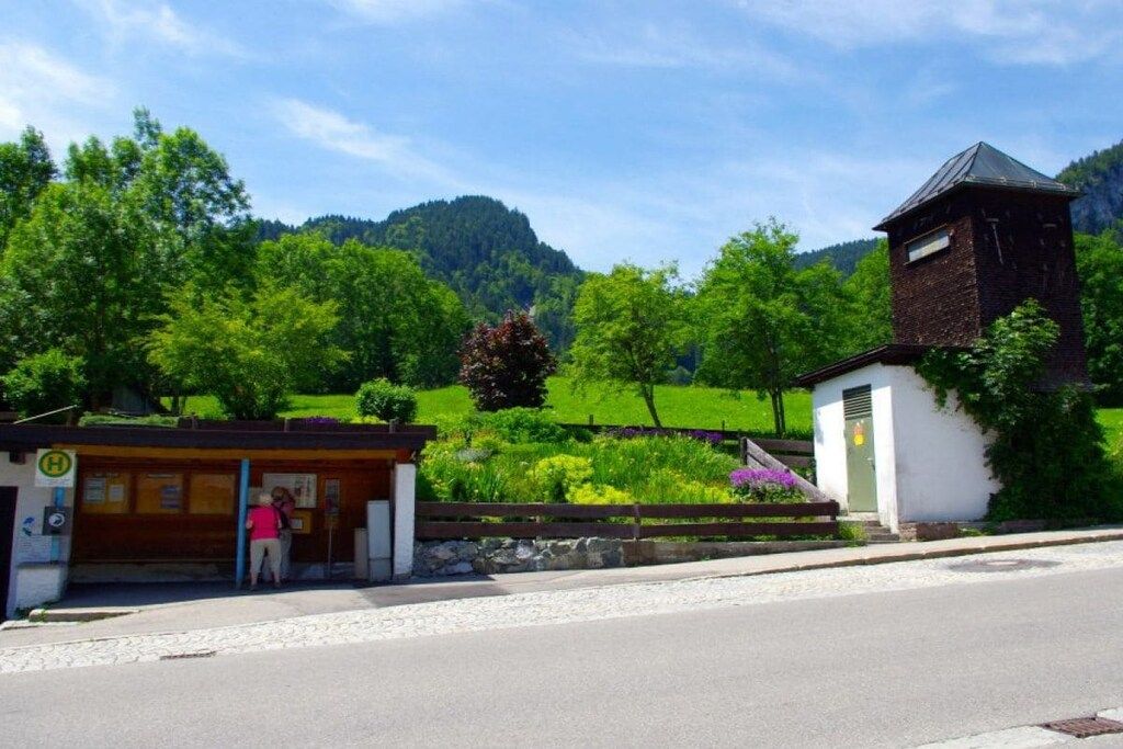 A rural setting with a small building that appears to be a station or a small post office, surrounded by lush greenery and a hill in the background.