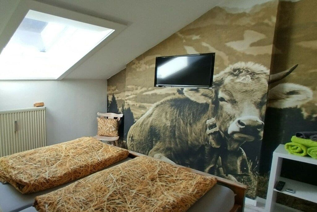 Une pièce dotée d’un téléviseur fixé au mur, d’une grande fresque murale représentant une vache, d’une lucarne et d’un lit recouvert de paille.