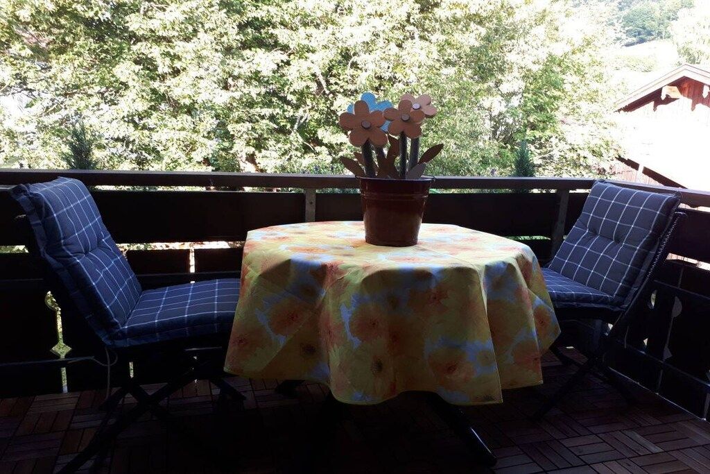A patio with a round table covered by a floral tablecloth, two blue cushioned chairs, and a potted plant with flowers.