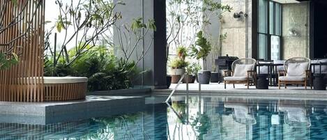 Área interna de piscina moderna, com água azul cristalina, cercada por espreguiçadeiras e plantas em vasos.