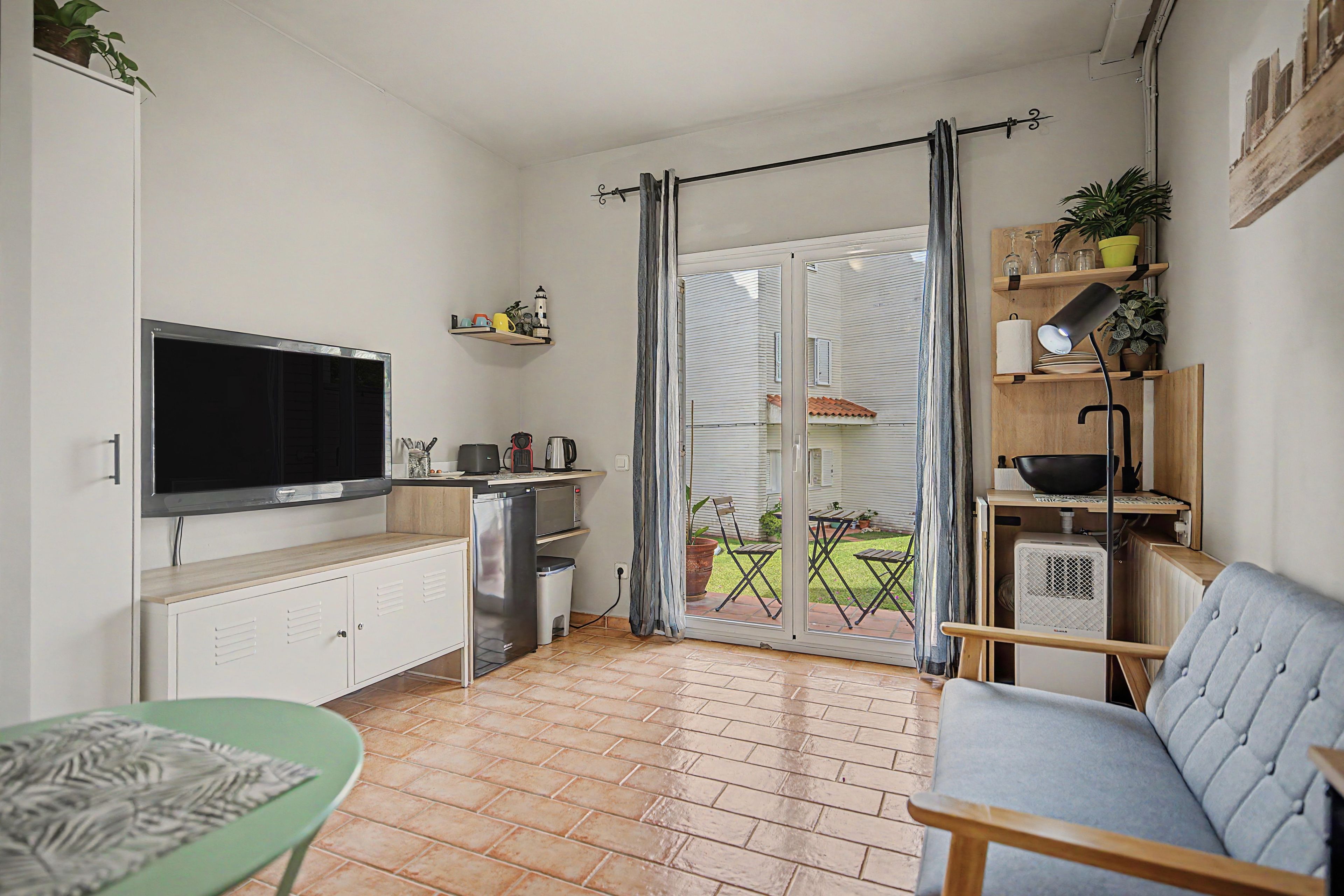 Een compacte leefruimte met een kitchenette, een flatscreen televisie, een eethoek met tafel en stoelen, en een schuifpui naar een terras.