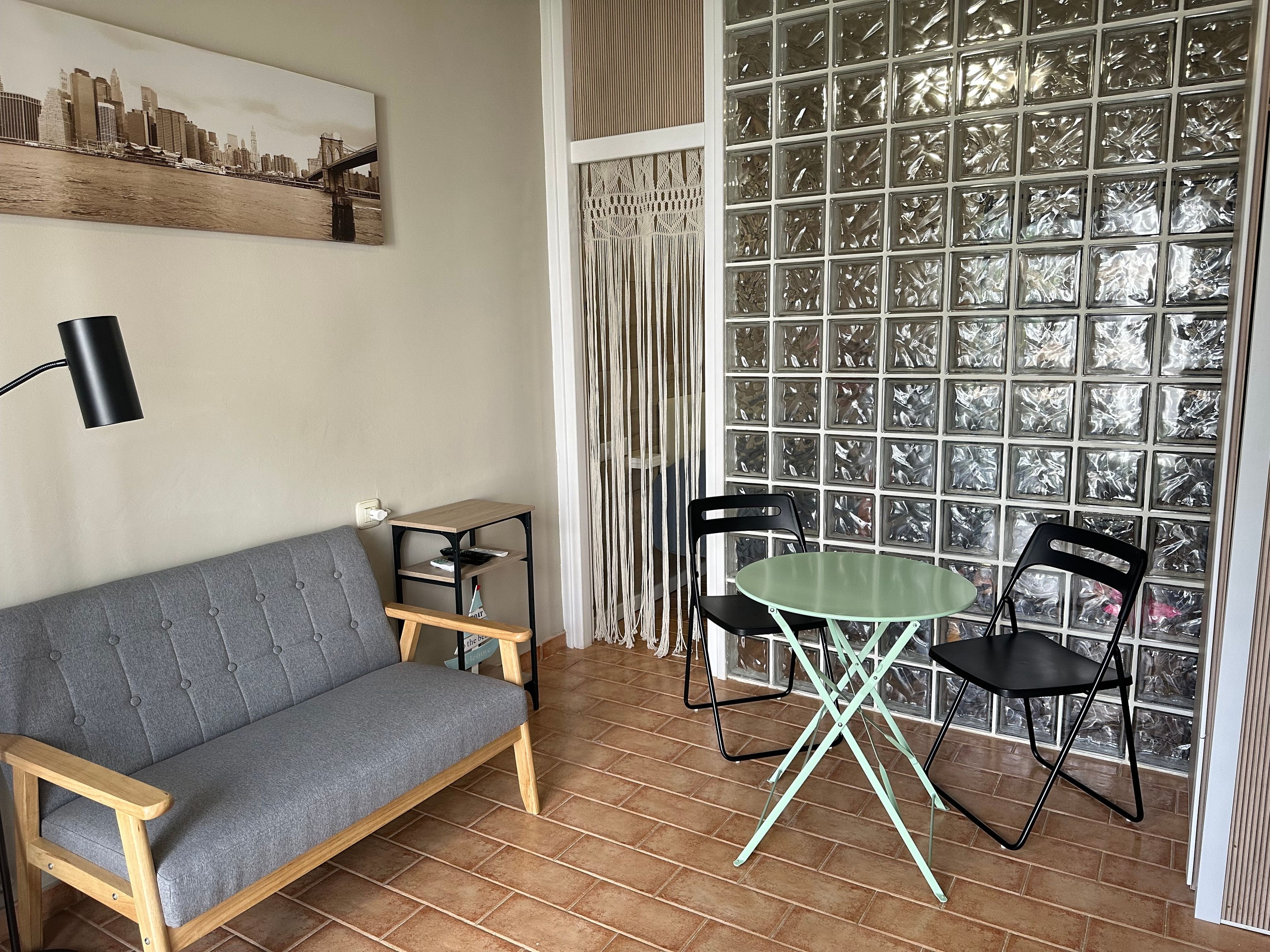 Une pièce avec un canapé gris, une petite table entourée de chaises, une lampe et un tableau représentant un paysage urbain accroché au mur.