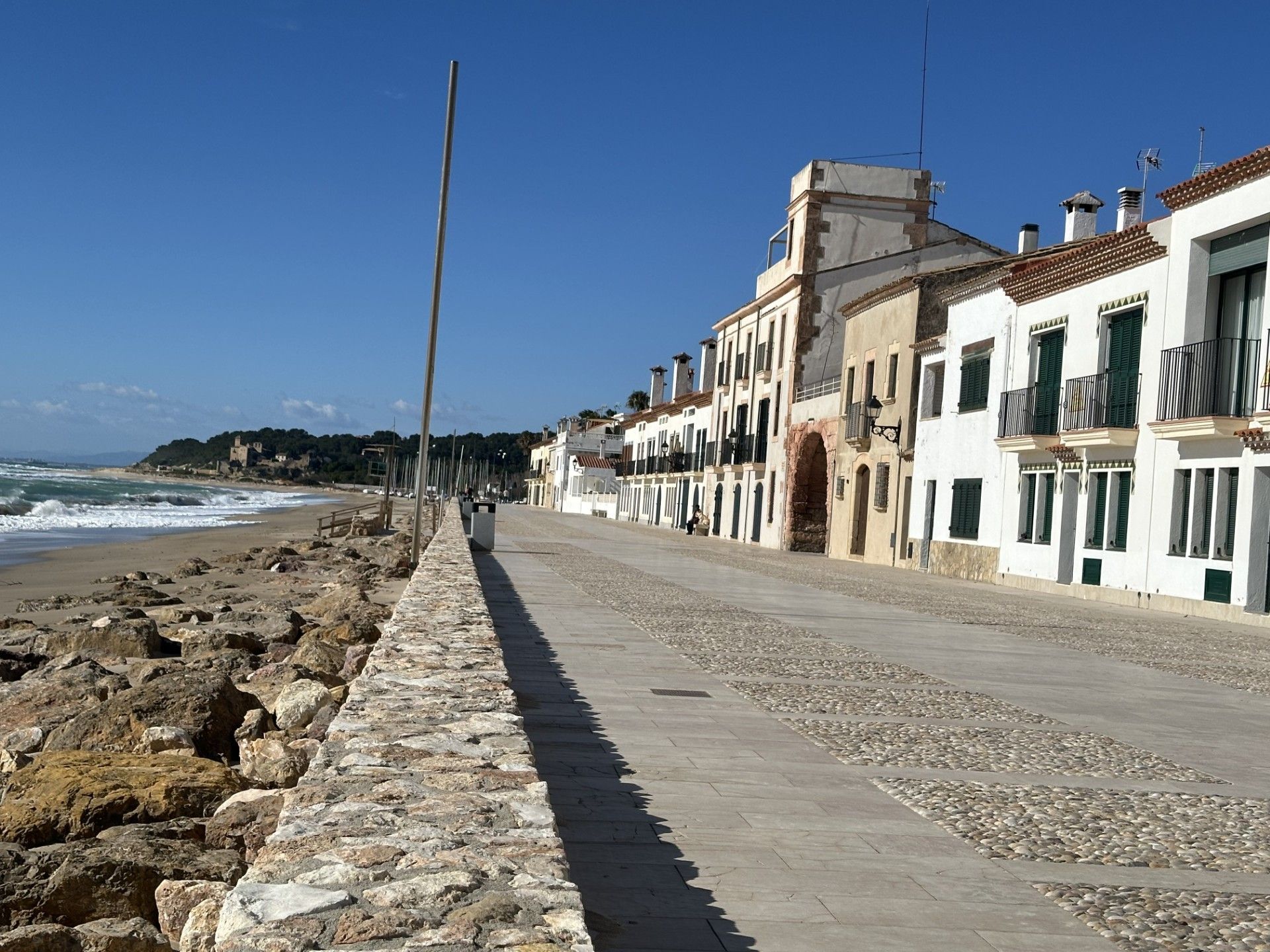 Une promenade en bord de mer, bordée de murs en pierre, de bâtiments dotés de balcons et surplombée d’un ciel dégagé.
