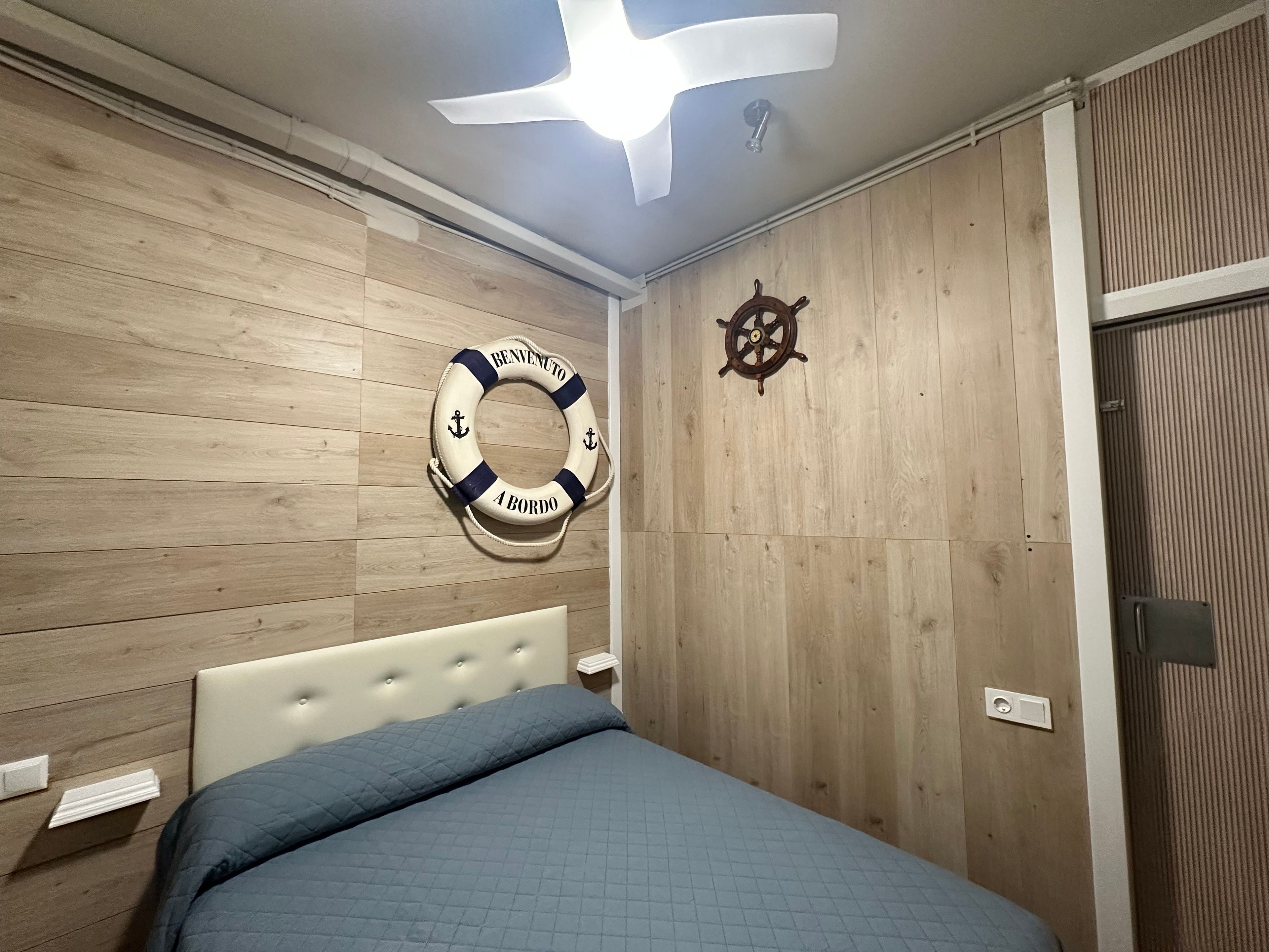 Une chambre à coucher avec un lit, des murs en bois, un ventilateur de plafond, une bouée de sauvetage et un décor représentant un gouvernail de navire.