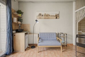 A compact living space with a kitchenette, a grey upholstered chair, a radiator, a floor lamp, and a cityscape painting on the wall.