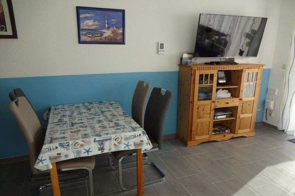 Un coin repas avec une table recouverte d’une nappe à motifs nautiques, deux chaises, un meuble en bois avec des étagères et un téléviseur fixé au mur.