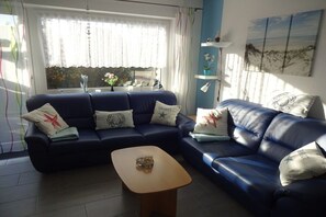 Ein Wohnzimmer mit einem blauen Sofagarnitur, einem Holztisch und einem Fenster mit transparenten Vorhängen.
