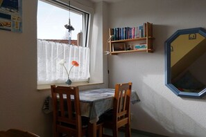 Ein Zimmer mit Esstisch und Stühlen, einem Bücherregal, einem Fenster mit Vorhang und einem Spiegel an der Wand.