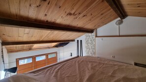 Una stanza con soffitto spiovente, travi in legno, un letto con lenzuolo bianco, una finestra con persiane di legno e una parete con carta da parati a motivi floreali.