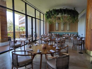Uma sala de jantar ampla, com mesas redondas e cadeiras de vime, grandes janelas e um teto com plantas pendentes.