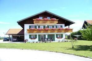 A two-story building with a sloped roof, balconies with flower boxes, and a small outdoor seating area. - Holiday apartment with 1 bedroom (Bad Bayersoien)