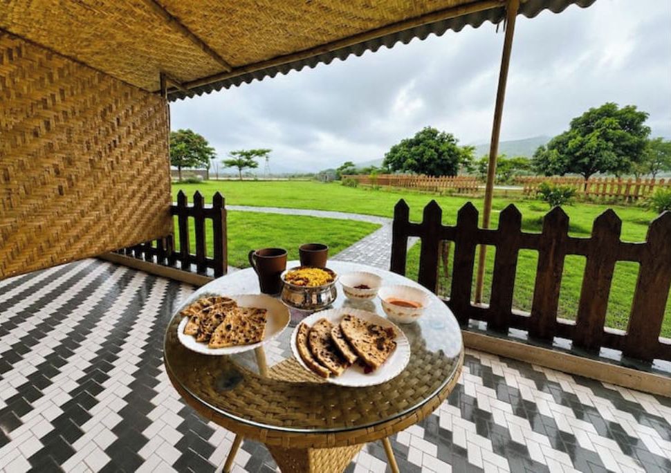 Une table ronde, dressée avec des plats et des boissons, se trouve sur une terrasse carrelée, d’où l’on peut admirer un champ verdoyant et des arbres.
