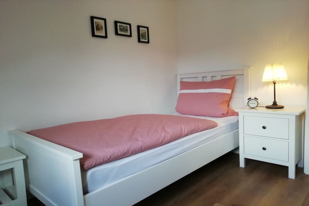 Una cama individual con estructura blanca y ropa de cama rosa, una mesita de noche con lámpara y un pequeño reloj de noche.
