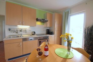 A kitchen with wooden cabinets, a stove, a sink, and a dining table with a bottle and glasses. - Dune grass with balcony (Dahme)