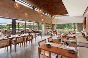 A spacious dining area with wooden tables and chairs, large windows, and hanging pendant lights. - Woods At Sasan (Mendarda)
