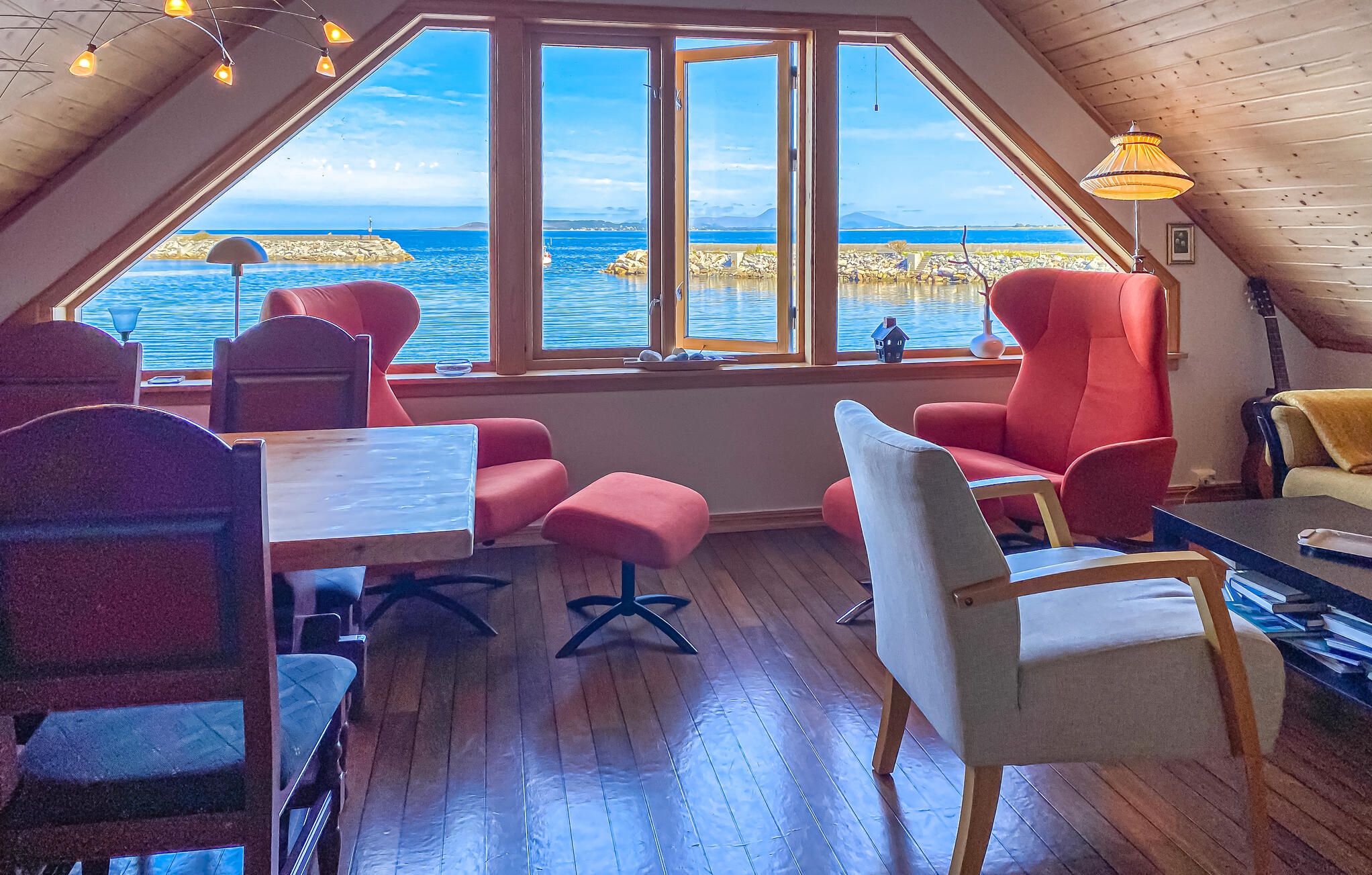 A cozy room with a large window overlooking a body of water, wooden flooring, and red and white furniture.