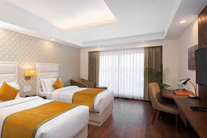 A hotel room with two beds, a desk, a chair, a TV, and a large window with curtains. - Cygnett Inn Paras Trademark by Wyndham (Dehradun)