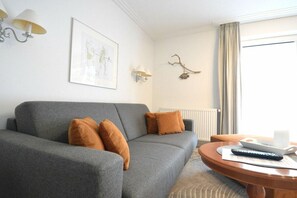 Un salon avec un canapé gris, des coussins marron, une table basse ronde en bois, un tableau encadré au mur et un radiateur.