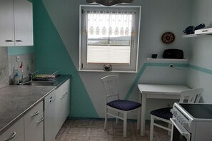 Eine kleine Küche mit Fenster, einem Waschbecken, einem Stuhl, einem Tisch und einer Mikrowelle.