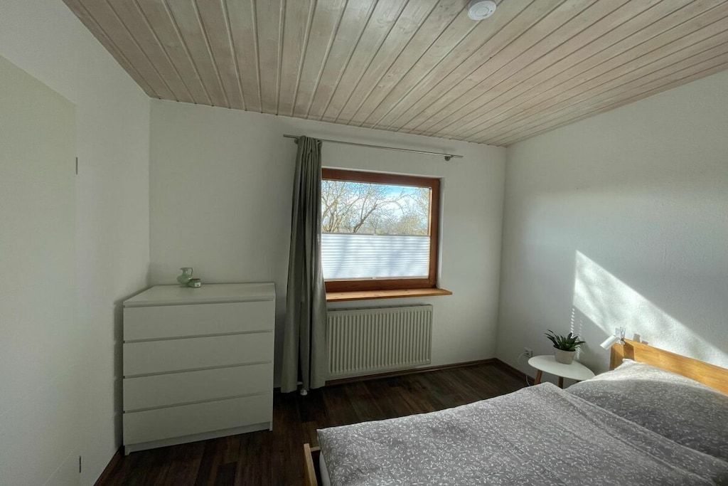 Une chambre à coucher avec un lit, une commode, une fenêtre avec des rideaux, un radiateur et une petite table sur laquelle est posée une plante.
