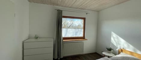 Un dormitorio con cama, cómoda, ventana con cortinas, radiador y una mesita con planta.